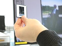 Eine Person hält ein fotografisches Dia vor einem Computermonitor, der ein gescanntes Bild anzeigt. Auf dem Bildschirm in einem hellen, modernen Arbeitsbereich ist eine Bildbearbeitungssoftware geöffnet.